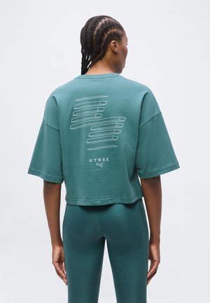 Person med fletninger iført en teal oversized cropped t-shirt med hvide grafiske linjer og tekst "HYROX" samt Puma-logo på ryggen.