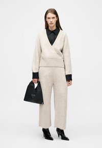 Femme portant un pull en maille beige ceinturé et un pantalon large assorti, chemise noire, bottines noires à bout pointu, tenant un sac à main noir.