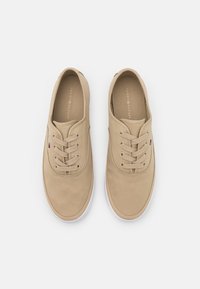 Par de zapatillas de lona beige con cordones y suelas blancas, pequeño detalle de la bandera de Tommy Hilfiger en los laterales, vistas desde arriba sobre un fondo blanco.