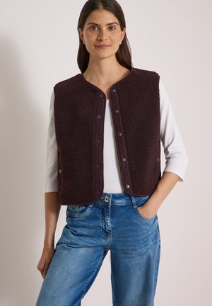 Frau mit langen dunklen Haaren, die ein weißes Top, blaue Jeans und eine dunkelbraune, geknöpfte Fleeceweste trägt, steht mit einer Hand in der Tasche.