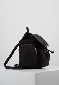 Czarny plecak z materiału z klapą, wieloma kieszeniami na zamek, złotym okuciem, regulowanym paskiem i gładką teksturą.