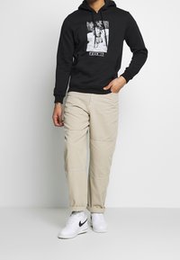 Černá mikina s kapucí s grafickým potiskem a textem, doplněná béžovými kalhotami. Bílá sportovní obuv celý outfit dokončuje.