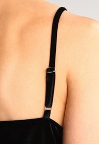 Musta samettinen hihaton paita hopeisella metallisella säätimellä, lepäämässä sileällä, rusketetulla ihon pinnalla.