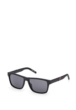 Occhiali da sole rettangolari neri con lenti scure, montatura opaca e sottili accenti rossi e blu sulle astine. Presentano linee dritte e un design pulito.