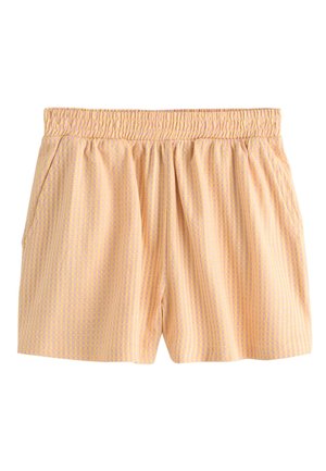 Kvinde med krøllet hår iført en ferskenfarvet stropløs top og matchende shorts, der står på en træbro ved havet.