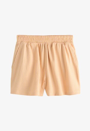 Donna con capelli ricci che indossa un top senza spalline color pesca e pantaloncini coordinati, in piedi su una passerella di legno vicino al mare.