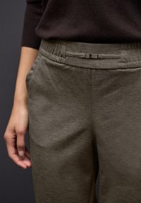 Olivgröna byxor med en texturerad tyg, elastisk midjeband och en liten metallspänne detalj. Fickor syns på sidorna.