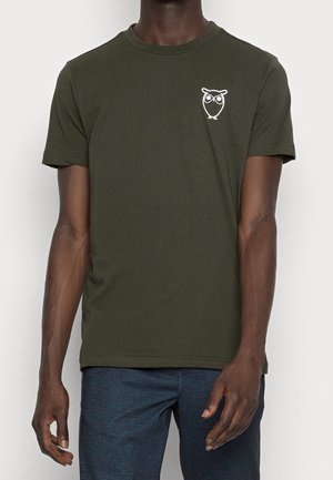 Mand iført en mørkegrøn t-shirt med en lille hvid uglelogo på venstre bryst og blå teksturerede bukser.