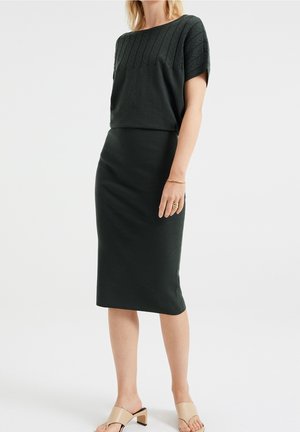 Femme portant un haut en maille noir à manches courtes et une jupe assortie jusqu'aux genoux, avec des sandales à talons beiges et des bijoux en or minimalistes.