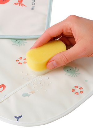 Hand hält einen gelben Schwamm und wischt Wassertropfen von einem weißen Lätzchen mit roten und grünen Mustern von Meerestieren und Rettungsringen ab.