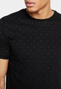 La camiseta de manga corta negra presenta un sutil patrón en todo su diseño con formas de lágrima en rojo oscuro, con cuello redondo y una textura suave.