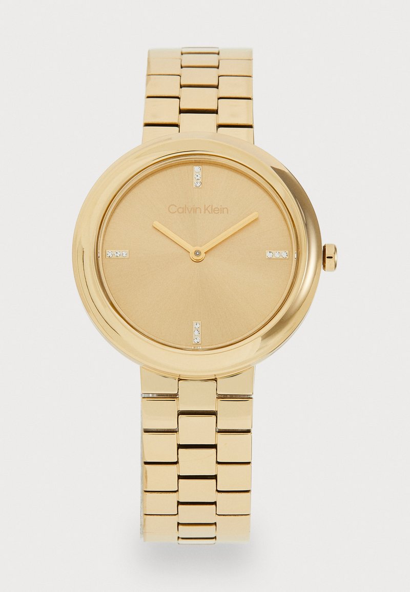 Gold-tone analog wristwatch with a smooth circular case, a matte gold dial, quartz movement, and five crystal hour markers on a metal bracelet.