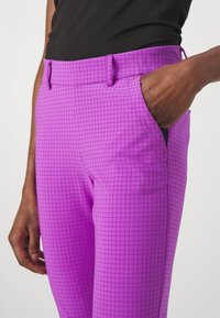 Person wearing purple checkered pants with one hand in pocket and black top against a white background.