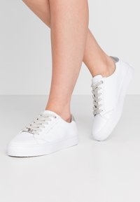 Legs wearing white low-top sneakers with gray laces and silver heel detail, standing on a light gray floor against a plain white background.