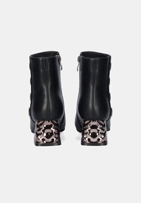 Botas de tobillo de cuero negro con acentos decorativos de botones y un tacón metálico con diseño. Cuenta con un zipper lateral para facilitar su uso.