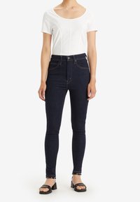 Jeans de talle alto de color azul oscuro con una silueta ajustada y costuras visibles, combinados con una camiseta blanca de manga corta y sandalias negras.