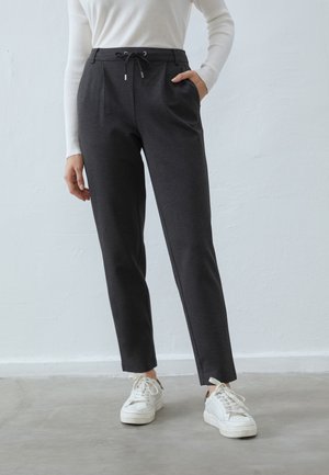 Osoba nosząca ciemnoszare spodnie z sznurkiem, biały długi rękaw oraz białe trampki, stojąca na szarej podłodze przed prostą ścianą.