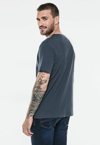 Grå bomullströja med korta ärmar och rund halsringning, visad från baksidan. Har synliga tatueringar på armen och avslappnad passform.