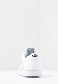 Baskets blanches Nike vues de dos, affichant le logo Nike noir sur le talon et une semelle blanche sur un fond blanc uni.
