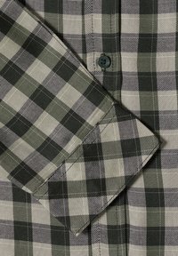 Gros plan d'une manche de chemise à carreaux verts et noirs avec poignet boutonné, coutures visibles et texture du tissu.
