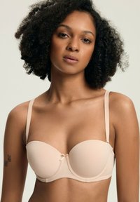 Kobieta z kręconymi włosami nosząca beżowy biustonosz bez ramiączek z cienkimi regulowanymi paskami, stojąca na jednolitym jasnym tle.