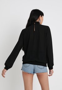 Blouse noire à manches longues, col froncé et poignets élastiques, associée à un short en denim clair, dévoilant un détail en forme de lucarne dans le dos.