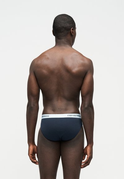Μυώδης άνδρας με σκούρο δέρμα, ντυμένος με μπλε εσώρουχα navy με λευκή ζώνη Emporio Armani, στέκεται με την πλάτη σε απλό ανοιχτό φόντο.
