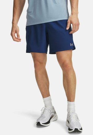 Man draagt marineblauwe sportshorts, witte sokken en witte hardloopschoenen, staat met ontspannen armen.