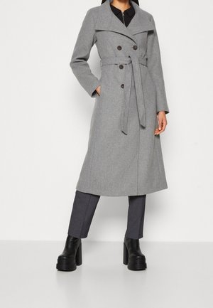 Kobieta w szarym dwurzędowym płaszczu z paskiem, ciemnych spodniach i masywnych czarnych butach na platformie, stojąca na jednolitym tle.