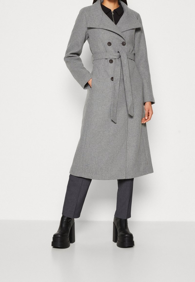 Femme portant un manteau gris ceinturé à double boutonnage, un pantalon foncé et des bottes plateformes noires épaisses, debout devant un fond uni.