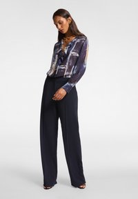 Langemouw blouse met ruches bij de hals in een abstracte print in paars, blauw en creme, gecombineerd met op maat gemaakte marineblauwe wijde broekspijpen.