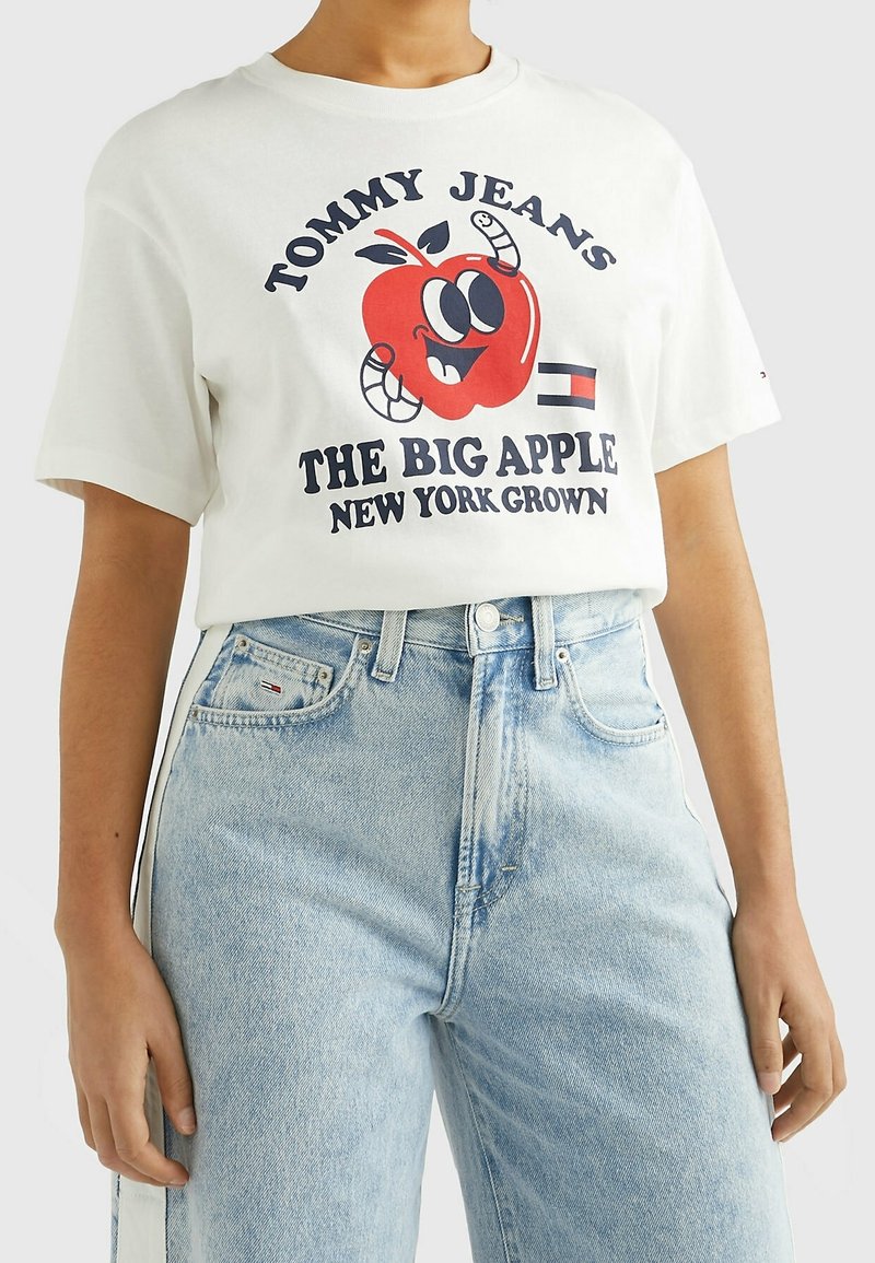 Bílý bavlněný tričko s grafikou červené usmívající se jablka. Text zní "TOMMY JEANS THE BIG APPLE NEW YORK GROWN." Uvolněný střih, krátké rukávy.