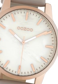 Rose gold watch featuring a white mother-of-pearl dial, minimalist hour markers, and a beige rubber strap. Logo positioned at the top.