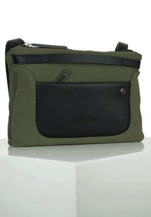 Oliwkowa torba na ramię z czarną przednią kieszenią, zamkiem błyskawicznym i regulowanym paskiem, spoczywająca na białej powierzchni.