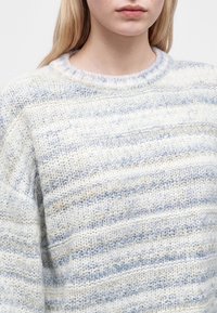 Jeune personne portant un pull en maille douce avec des rayures horizontales bleu clair, blanc et beige.
