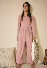 Set di pigiama in morbido tessuto a costine rosa chiaro, composto da una maglietta a maniche lunghe e pantaloni larghi con vita elastica, abbinato a un cuscino con motivo floreale.