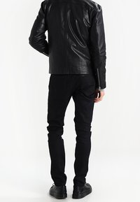 Schwarze Lederjacke mit taillierter Passform, sichtbaren Nähten und Reißverschlüssen. Kombiniert mit schwarzen schmal geschnittenen Jeans und schwarzen Schuhen.