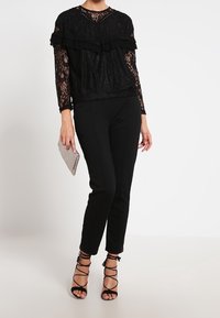 Haut en dentelle noire avec des couches volantes, associé à un pantalon noir ajusté et des talons noirs à lanières. Tenant une pochette en argent texturé.