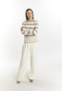 Gestreifter Rippstrickpullover in Beige und Weiß, kombiniert mit lockeren weißen Hosen. Das Model steht mit verschränkten Armen vor einem neutralen Hintergrund.