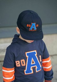 Laajentuneen merivoimien baseball-lakki, jossa on sininen ja oranssi brodeerattu teksti; yhteensopiva laivaston sininen collegepaita, jossa on suuri sininen kirjain "A" ja oranssit raidat hihoissa.