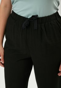 Pantaloni neri elasticizzati con cintura regolabile, caratterizzati da una texture liscia e una vestibilità comoda, presentati su uno sfondo chiaro.
