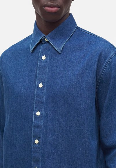 Homme portant une chemise en jean bleue boutonnée avec un col classique, montré de la poitrine vers le haut sur un fond uni.