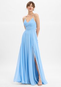 Frau in einem hellblauen, bodenlangen, ärmellosen Kleid mit Spaghettiträgern und einem seitlichen Schlitz bis zum Oberschenkel, stehend vor einem schlichten weißen Hintergrund.