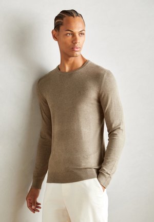 Pull léger marron clair avec un col rond et des poignets et un ourlet côtelés, fabriqué en tissu tricot doux. Le modèle porte un pantalon blanc.