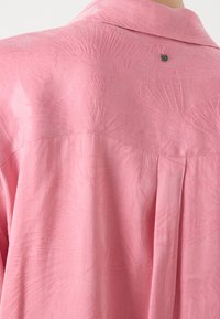 Vue arrière d'une chemise rose avec un motif jacquard floral subtil et une petite étiquette métallique ronde sous le col.