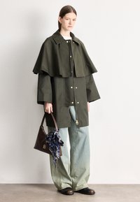 Casaco oversized de poli-algodão em verde-oliva com design de capa rodada, acessórios dourados, combinado com jeans de pernas largas azul claro e uma mala castanha.