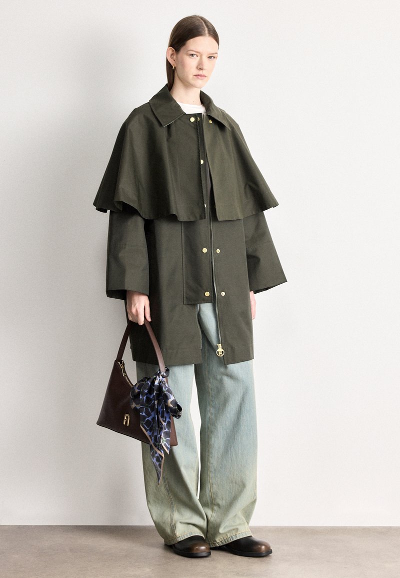 Casaco oversized de poli-algodão em verde-oliva com design de capa rodada, acessórios dourados, combinado com jeans de pernas largas azul claro e uma mala castanha.