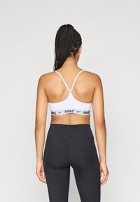 Bela športna nedrček z nastavljivimi tankimi naramnicami in črnim trakom z logotipom Nike, kombiniran z visokim pasom črnih pajkic, gladka tekstura tkanine.
