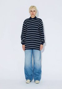 Maglietta polo a maniche lunghe a righe blu navy e bianche abbinata a jeans larghi azzurri chiaro. Il modello è in piedi davanti a uno sfondo bianco semplice.