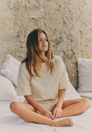 Hosszú hajú nő keresztbe tett lábbal ül egy fehér ágyon, mögötte egy texturált bézs kőfal, világos bézs inget és rövidnadrágot visel.